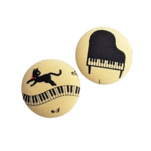 Könnte beinhalten: Zwei runde, beige Stoffknöpfe. Ein Knopf zeigt eine schwarze Katze, die über einer Klaviertastatur rennt. Der andere Knopf hat eine Illustration eines schwarzen Flügels. Musik-Themen-Knöpfe.