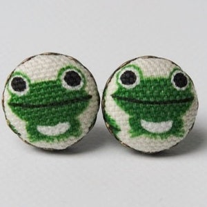 May include: A pair of green frog-themed fabric earrings. The earrings are round and have a white background with a green frog face in the center. The frog has black eyes and a white smile.