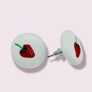 Peut inclure: Une paire de boucles d'oreilles clous en tissu blanc avec un motif de fraise brodé en rouge.