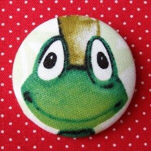 Op de afbeelding: Een groene kikker met een bruine kroon op zijn kop. De kikker heeft grote witte ogen met zwarte pupillen. De kikker is gemaakt van stof en heeft een gestikte glimlach.