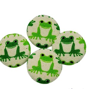 Puede incluir: Cuatro botones verdes de tela con forma de rana sobre fondo blanco. Las ranas sonríen y tienen ojos negros. Los botones tienen una superficie texturizada.