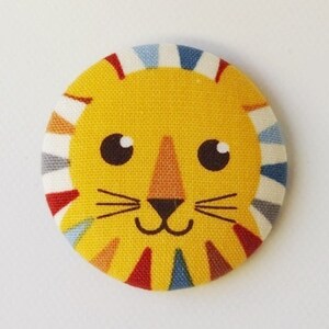 Peut inclure: Un bouton en tissu jaune avec un motif de visage de lion. Le lion a un nez noir, des moustaches noires et des yeux bleus. Le bouton est entouré d'une bordure rayée colorée.