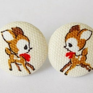 May include: A pair of white fabric button earrings with a brown and white embroidered deer design. Each deer has a red bow.