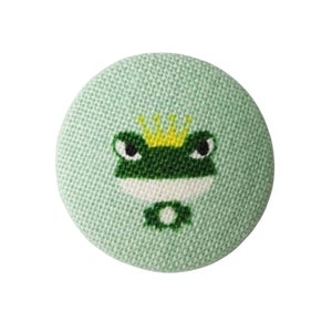 Könnte beinhalten: Ein runder, hellgrüner Knopf mit einem Froschdesign. Der Frosch ist grün und weiß und trägt eine goldene Krone. Der Knopf hat eine strukturierte Stoffoptik.