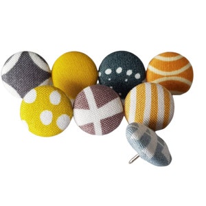 Könnte beinhalten: Ein Satz von acht stoffbezogenen Stecknadeln mit verschiedenen geometrischen Mustern in Grautönen, Gelb und Weiß.