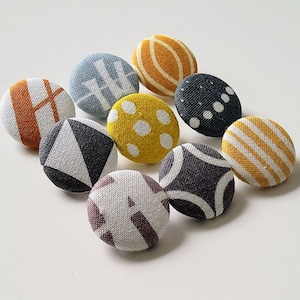 Può includere: Assortimento di nove bottoni rivestiti in tessuto con vari motivi geometrici e astratti in tonalità di giallo, arancione, grigio e blu.
