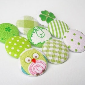 Könnte beinhalten: Eine Sammlung dekorativer Stoffknöpfe in Grün- und Weißtönen. Die Knöpfe zeigen Muster wie Punkte, Streifen, Vichy-Karo und ein vierblättriges Kleeblatt. Ein Knopf hat ein Eulen-Design, ein anderer ein Schaf.