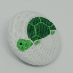 Peut inclure: Un bouton en tissu blanc avec un motif de tortue verte. La tortue a une carapace verte avec un œil noir.