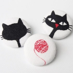May include: Three white fabric buttons with black cat designs. One button features a black cat with a red nose, another button features a black cat silhouette, and the third button features a red ball of yarn.