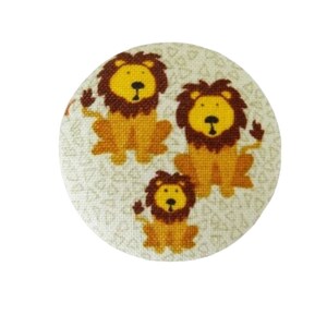Peut inclure: Un bouton rond avec un fond blanc et un motif de petits cœurs. Trois lions sont représentés sur le bouton, un grand et deux plus petits, tous avec des crinières brunes et des corps jaunes.