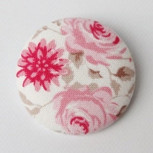 Può includere: Un bottone rotondo in tessuto bianco con un motivo floreale rosa e rosso. Il bottone presenta una grande rosa rosa con un fiore rosso più piccolo sullo sfondo.