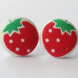 Może przedstawiać: Dwa okrągłe guziki z tkaniny z wzorem truskawki. Każdy guzik przedstawia czerwoną truskawkę z białymi nasionami i zielonym liściem na białym tle. Idealne do projektów krawieckich.