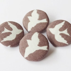 Puede incluir: Cuatro botones de tela marrón con siluetas de pájaros blancas. Los botones tienen un diámetro de aproximadamente 2,5 cm.