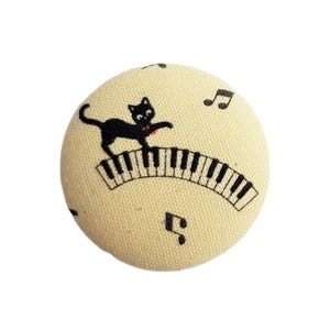 Peut inclure: Un bouton en tissu beige avec un chat noir jouant du piano avec des notes de musique imprimées autour.
