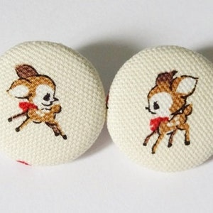 Op de afbeelding: Twee witte stoffen knopen met een bruin en wit cartoon hert ontwerp. Elk hert draagt een rode strik.
