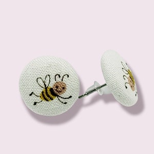 Può includere: Un paio di orecchini a bottone in tessuto bianco con un design di ape cartoon. L'ape è gialla e nera con la testa e le ali marroni.