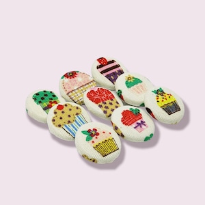 Pode incluir: Conjunto de oito botões de tecido em forma de cupcake em várias cores e designs. Os botões são redondos e apresentam diferentes designs de cupcake com cobertura, granulado e cerejas.