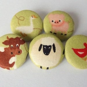 Könnte beinhalten: Fünf Stoffknöpfe mit verschiedenen Bauernhoftiermotiven. Die Knöpfe sind grün mit weißen, braunen, rosa und roten Tierillustrationen. Die Tiere umfassen eine Kuh, ein Schwein, ein Schaf, eine Gans und einen Hahn.