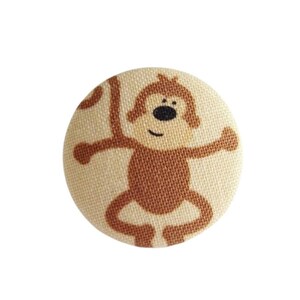 Peut inclure: Un bouton en tissu beige avec un motif de singe brun. Le singe a un visage souriant et tient ses bras tendus.