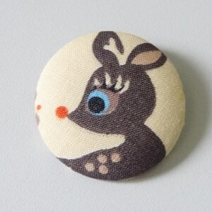 May include: A brown and white fabric button featuring a cartoon reindeer design. The reindeer has a blue eye, eyelashes, and a red nose.