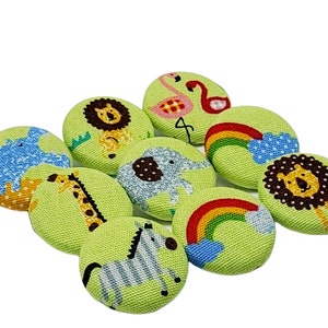 Op de afbeelding: Een set van negen groene stoffen knopen met verschillende dier- en regenboogontwerpen. De knopen tonen een leeuw, een zebra, een giraffe, een olifant, een flamingo en een regenboog.