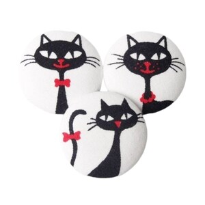 Puede incluir: Tres botones de tela blanca con diseños de gatos negros. Cada gato tiene un lazo o collar rojo.