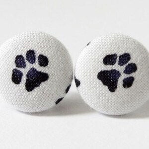 May include: A pair of white fabric button earrings with a black paw print design.
