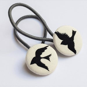 Könnte beinhalten: Zwei graue Haargummis mit weißen Stoffknöpfen, die schwarze Silhouetten von Vögeln zeigen.