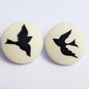 Op de afbeelding: Twee witte stoffen knopen met zwarte geborduurde vogelsilhouetten. De vogels kijken in tegengestelde richtingen.