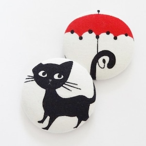 Könnte beinhalten: Zwei Stoffknöpfe mit weißem Hintergrund und schwarzen Umrissen. Ein Knopf zeigt eine schwarze Katze auf weißem Hintergrund. Der andere Knopf zeigt einen roten Regenschirm auf weißem Hintergrund.