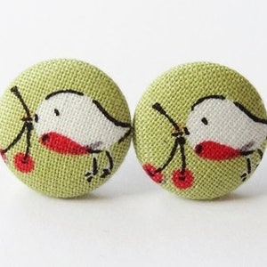 Könnte beinhalten: Ein Paar runde Knopf-Ohrringe mit hellgrünem Stoffhintergrund. Jeder Ohrring zeigt einen weißen Vogel mit rotem Schnabel und schwarzer Umrandung, der einen Zweig mit roten Kirschen hält. Ein einzigartiges Accessoire.