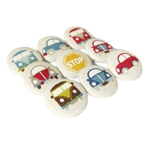 Può includere: Un set di dieci bottoni in tessuto con varie auto dei cartoni animati colorate e un cartello di stop giallo. I bottoni sono rotondi e hanno uno sfondo bianco.