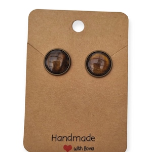 Può includere: Un paio di orecchini a bottone marroni e dorati con una finitura scintillante. Gli orecchini sono esposti su una carta marrone con il testo "Handmade with love".
