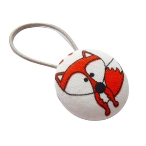 May include: A white fabric-covered hair tie featuring a cartoon fox design. The fox is depicted in orange and white with black accents. The hair tie has a gray elastic band.