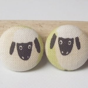Peut inclure: Deux boutons recouverts de tissu avec un motif de mouton. Les moutons sont blancs avec des visages et des oreilles noirs. Les boutons sont sur une surface en bois.