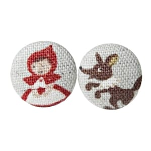 Könnte beinhalten: Zwei Stoffknöpfe mit einer rotgehüteten Mädchen und einem braunen Wolf. Die Knöpfe sind rund und haben einen weißen Hintergrund.