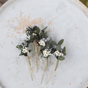 May include: Five gold hair pins with small white flowers and green leaves attached to the end of each pin. The pins are arranged on a white ceramic plate.
