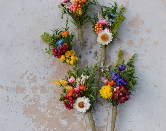 May include: A collection of colorful floral hair pins. Each pin features a small bouquet of dried flowers in shades of red, yellow, orange, pink, and white, with green foliage. The pins are arranged on a light gray surface.