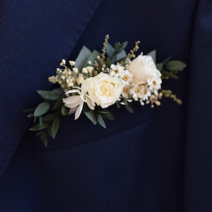 Op de afbeelding: Een witte roos en kleine witte bloemen met groene bladeren, gerangschikt in een corsage.