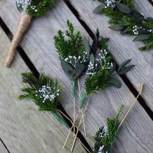Può includere: Una collezione di piccoli arrangiamenti floreali con rametti di verde e fiori bianchi. Gli arrangiamenti sono fissati con forcine dorate e sono pronti per essere utilizzati come accessori per capelli o boutonnières.