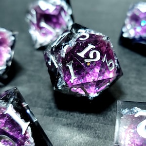 May include: A collection of handmade dice, featuring a blend of purple and clear resin with white numerals. The edges are highlighted with silver. The dice are arranged on a dark surface, emphasizing their distinctive design and craftsmanship.