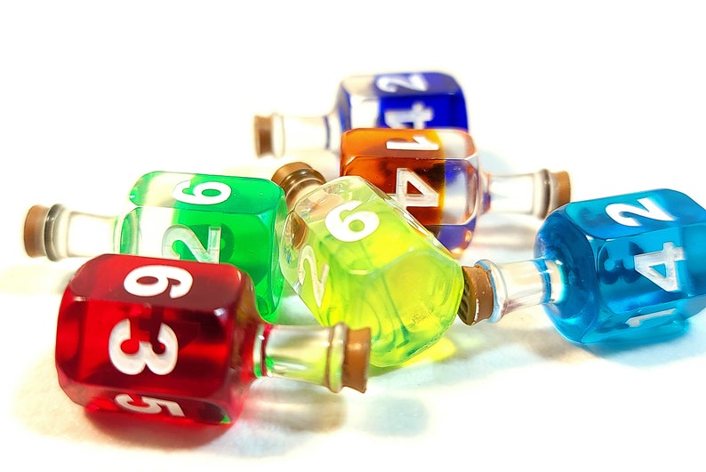 May include: Six colourful glass bottles with cork stoppers and white numbers on the sides. The bottles are filled with different coloured liquids and are lying on a white surface.