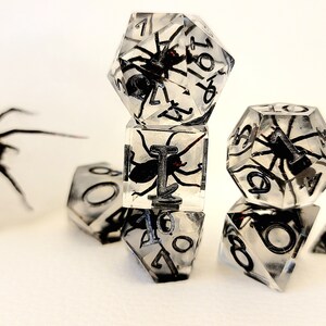 May include: A set of six clear dice with black numbers and a spider encased in each die. The dice are a d4, d6, d8, d10, d12, and d20.