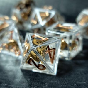 May include: A set of polyhedral dice made from clear resin, each containing a metallic gold-colored skeletal structure inside. The numbers on the faces of the dice are engraved and filled with a metallic gold-colored pigment for high visibility.