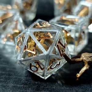 May include: A twenty-sided die made of clear resin with a brown, skeletal-like structure embedded inside. The numbers on the die are filled with a metallic copper-colored pigment. A small, brown, textured creature figure is positioned next to the die.