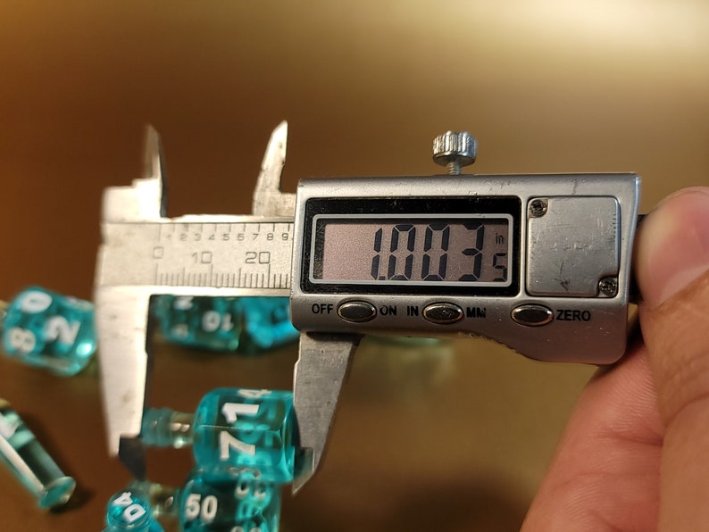以下が含まれることがあります： 1.003インチを測定する銀色のデジタルノギス。ノギスは、71という数字が書かれたターコイズブルーのプラスチック製のサイコロを測定しています。背景には、他のターコイズブルーのサイコロがあります。