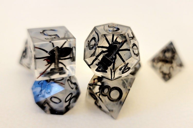 May include: A set of clear resin dice with a black spider encased inside each die. The dice are numbered 1 through 6 and are perfect for tabletop gaming.