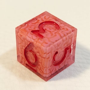 Op de afbeelding: Een doorzichtige roze dobbelsteen met rode cijfers, gevormd als een brein. De cijfers op de dobbelsteen zijn 2, 3, 4, 5 en 6.