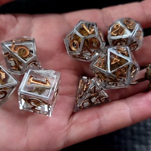 May include: A set of seven polyhedral dice made of clear resin with metallic gold numbers. Each die contains a small, detailed skeletal figure suspended inside the transparent material. The set includes various geometric shapes for tabletop gaming.