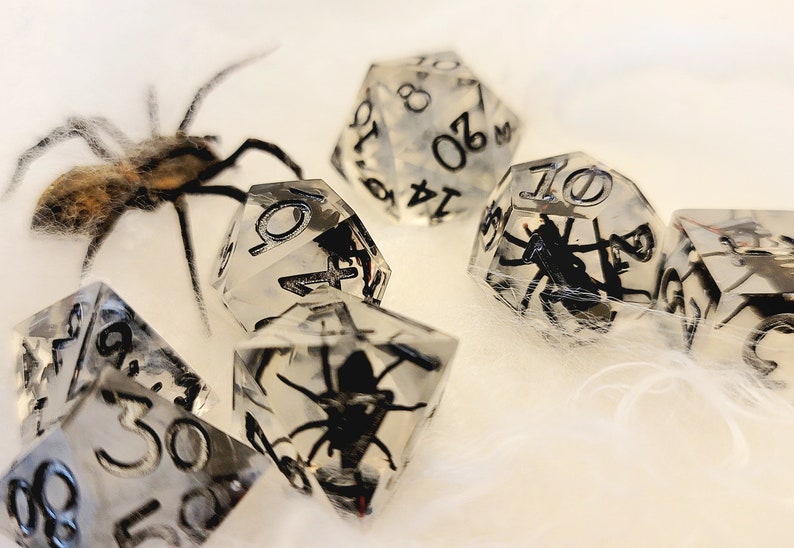 May include: A set of clear resin dice with black spiders encased inside. The dice are numbered 4, 6, 8, 10, and 20. The dice are on a white fuzzy surface.